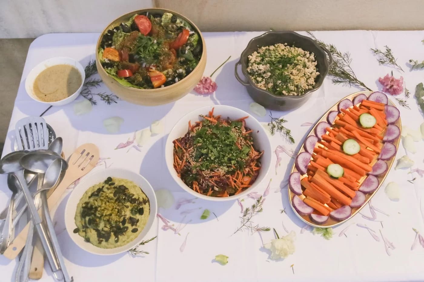 Un buffet végétal vu de dessus sur une nappe blanche : salades, taboulé, carottes râpées, crudités, houmous