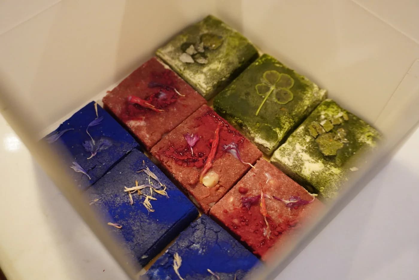 Carrés de chocolat cru bleu, rouge et vert, parés de pétales, dans leur coffret